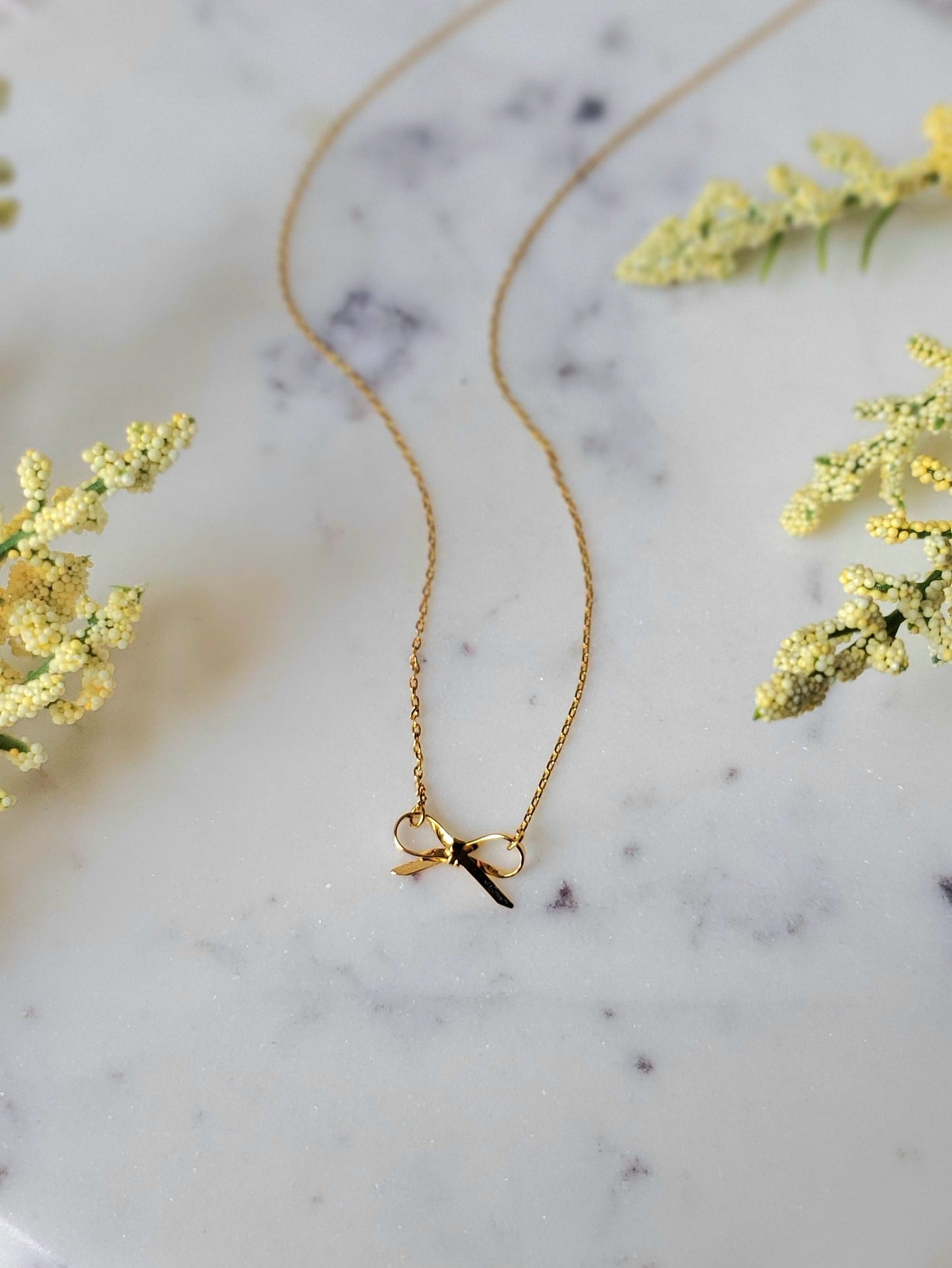 Gold necklace with a small pendant on a marble surface with yellow flowers.