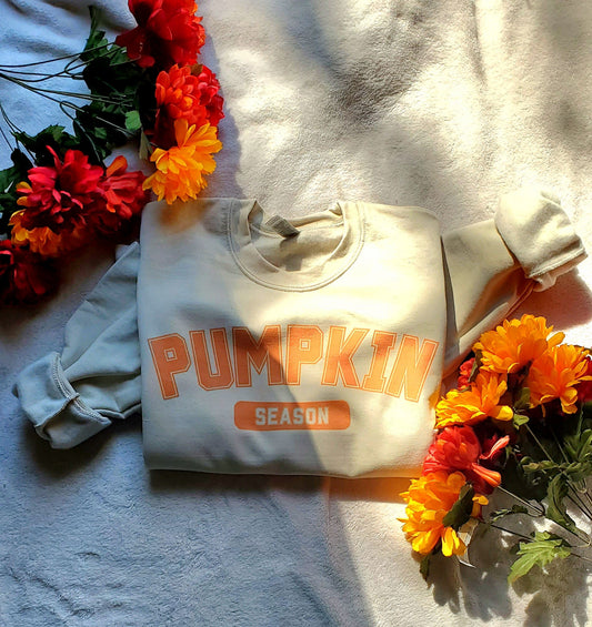 White sweatshirt with 'Pumpkin Season' text on a bed with floral decorations.